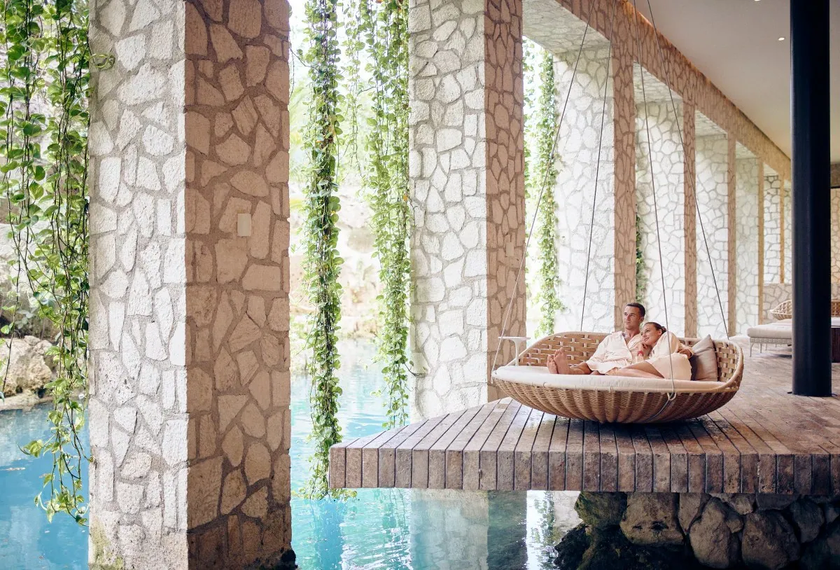 2 guests in a hanging nest above the water of the Muluk Spa, between stone columns and trailing plants