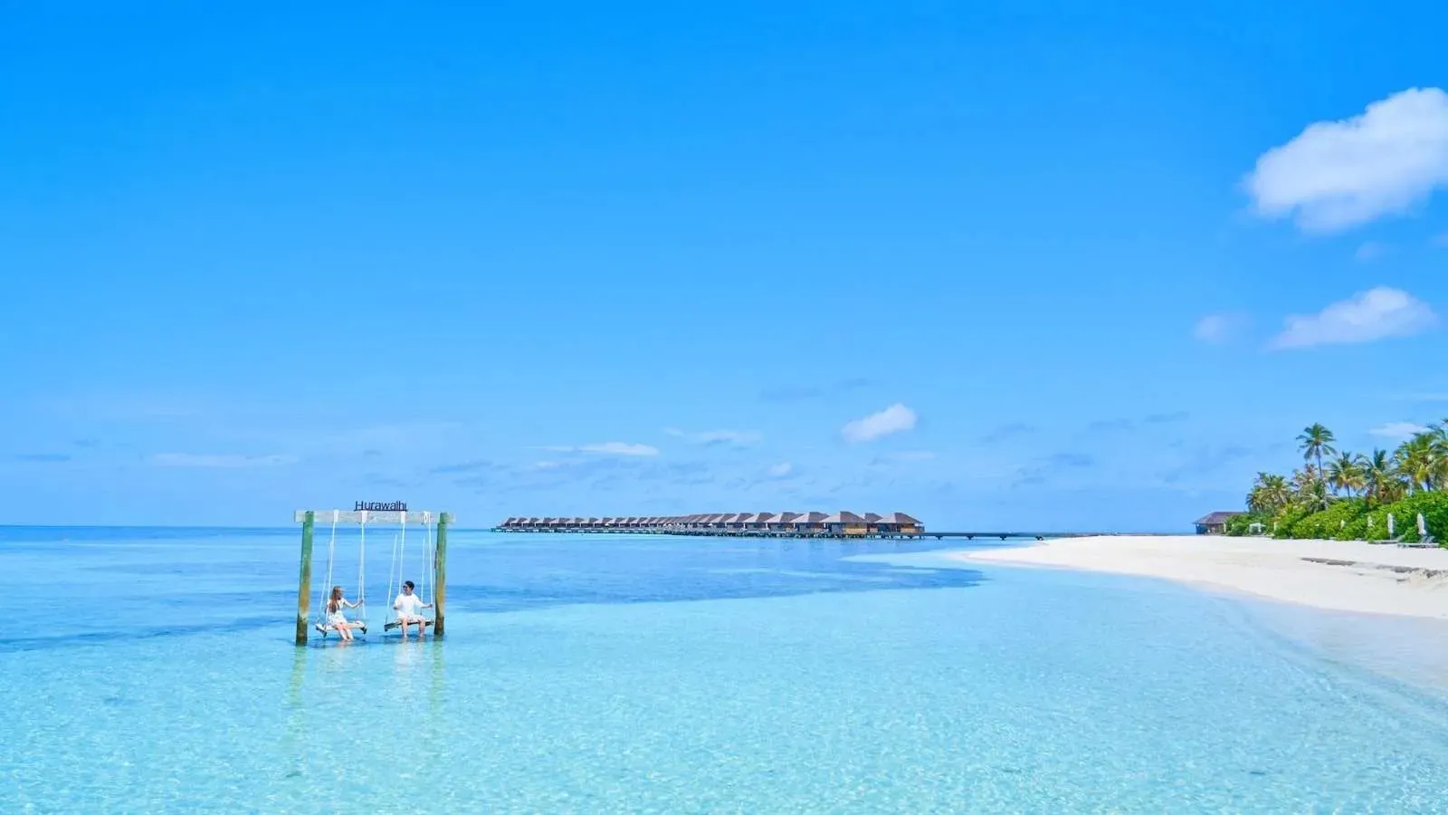 A swing in the shallow lagoon, the white beach and the overwater villas beyond