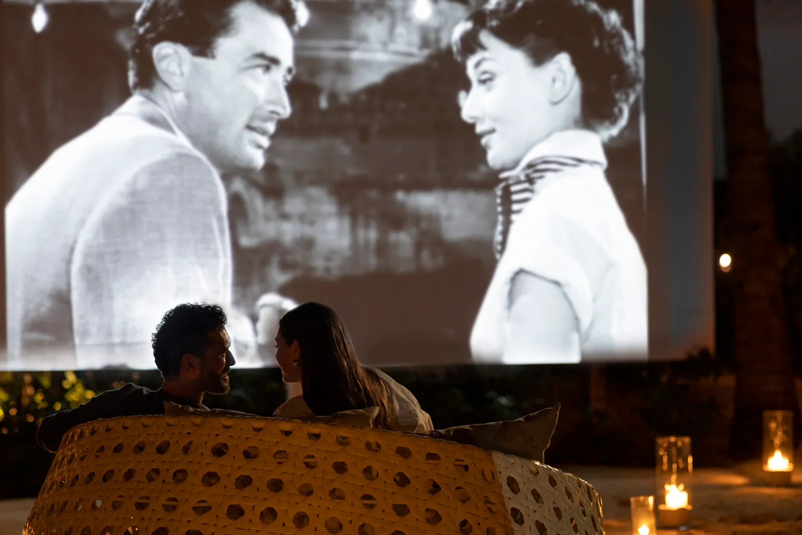 A black-and-white film projected outdoors, a couple on the cushions