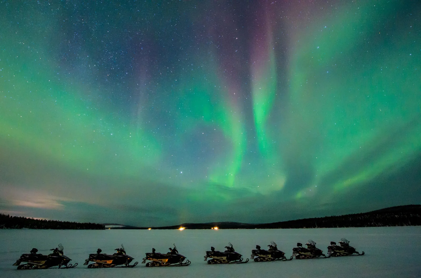 Green and violet northern lights fanning out above a frozen lake, with a line of snowmobiles standing on the snow.
