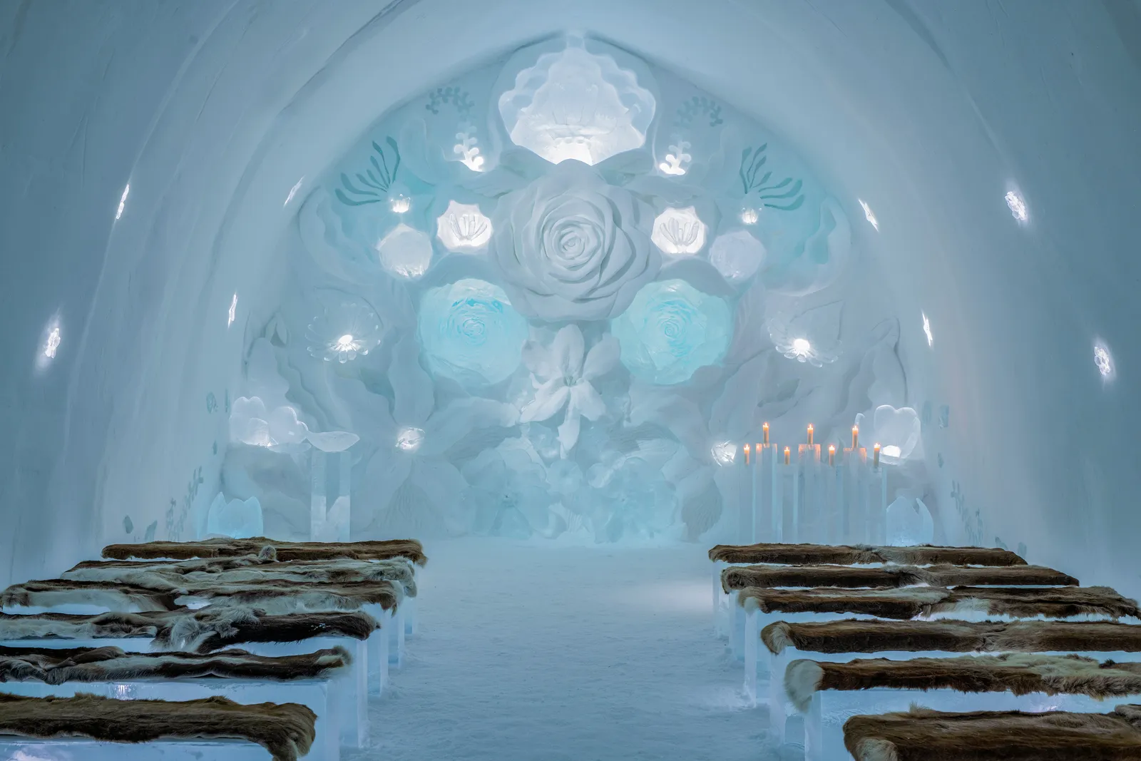 The Ceremony Hall Gaia’s Bloom, ICEHOTEL 35: rows of ice benches covered in reindeer hides facing a wall of flowers carved in snow, with ice candles to the right.