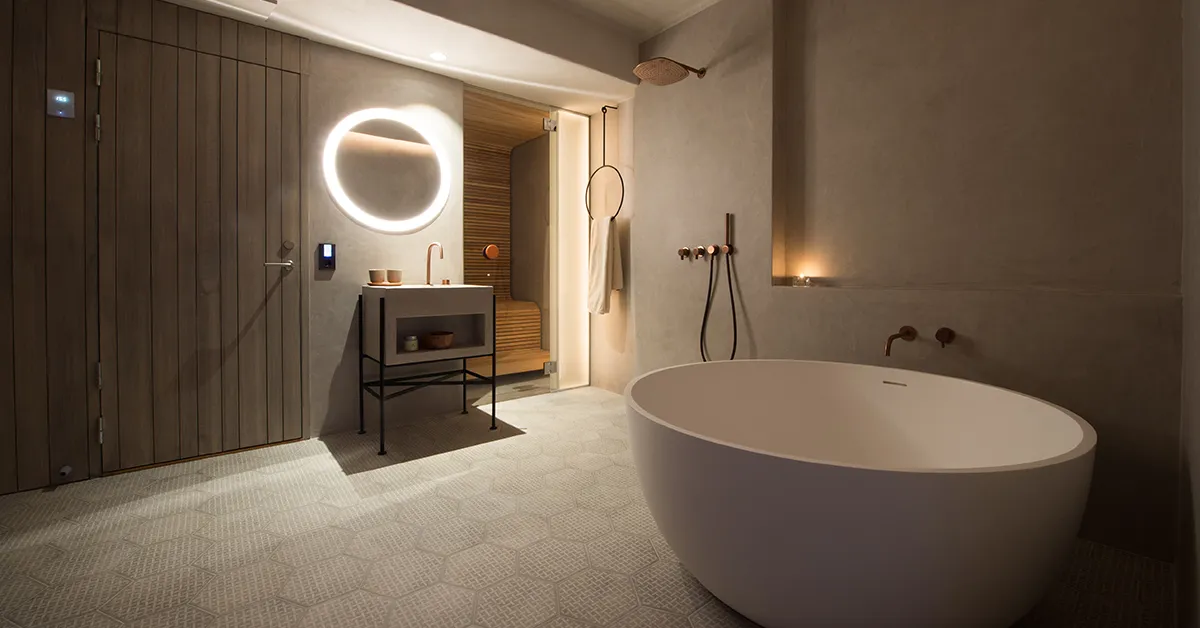 The heated bathroom of a Deluxe Suite: a freestanding oval bath, a round lit mirror above the basin, a rain shower and the glass door of the sauna, in grey stone and dark wood.