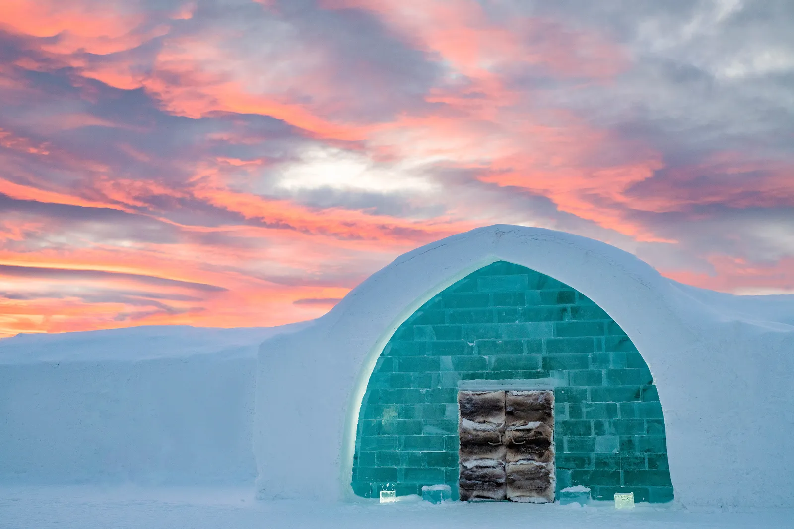 The entrance to the winter hotel: an arch of turquoise ice blocks set in a wall of snow, its doors covered in reindeer hide, under pink clouds.