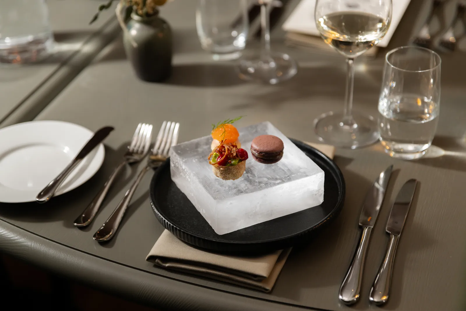 A table laid in the ICEHOTEL Restaurant: 3 small bites served on a block of clear ice set on a black plate, with cutlery, a glass of white wine and a glass of water.