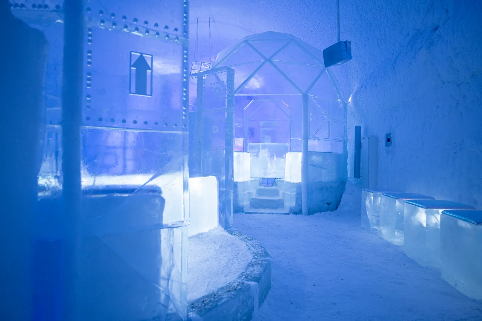 The ICEBAR in ICEHOTEL 365 in violet light: ice walls riveted like a spacecraft hull, a geodesic frame of ice and benches of ice blocks on the snow floor.