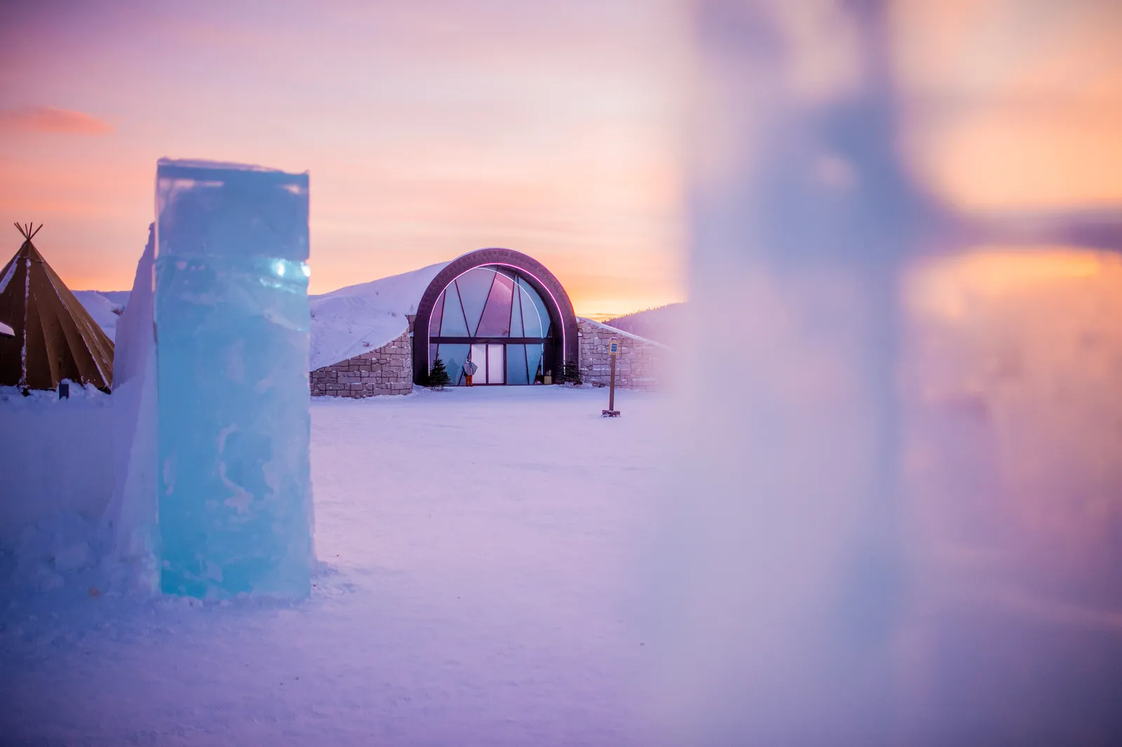 ICEHOTEL 365 at dusk in winter: the glazed arch of its entrance under a snow-covered roof, a tall block of clear ice in front and a Sámi tent to the left, in pink light.