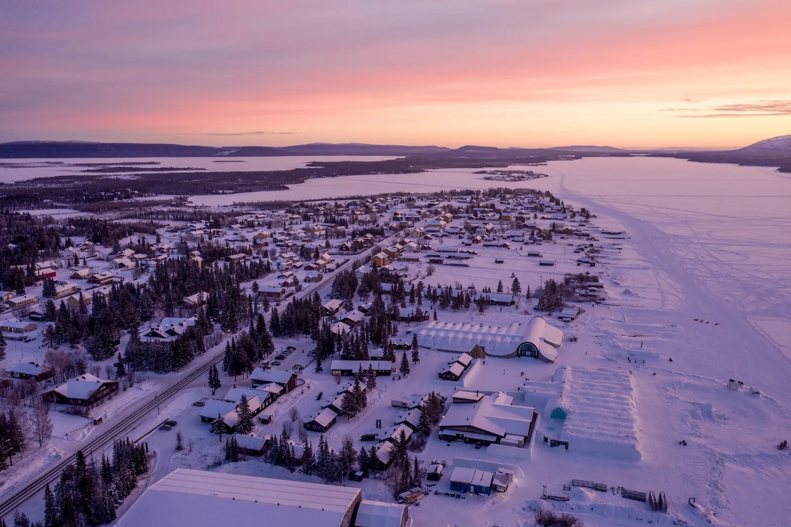 Jukkasjärvi from the air in winter: the village houses along the road, the snow-covered Torne River to the right and pink light on the horizon.