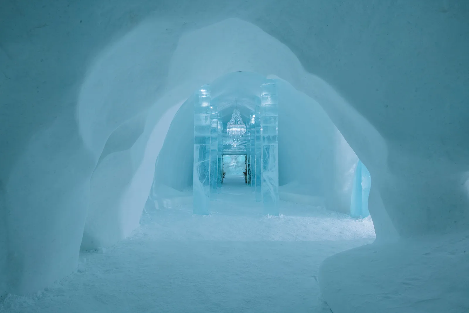 A vaulted corridor of snow in the winter hotel leading to a gateway of clear ice columns, an ice chandelier beyond: Main Hall Quasar, ICEHOTEL 35.