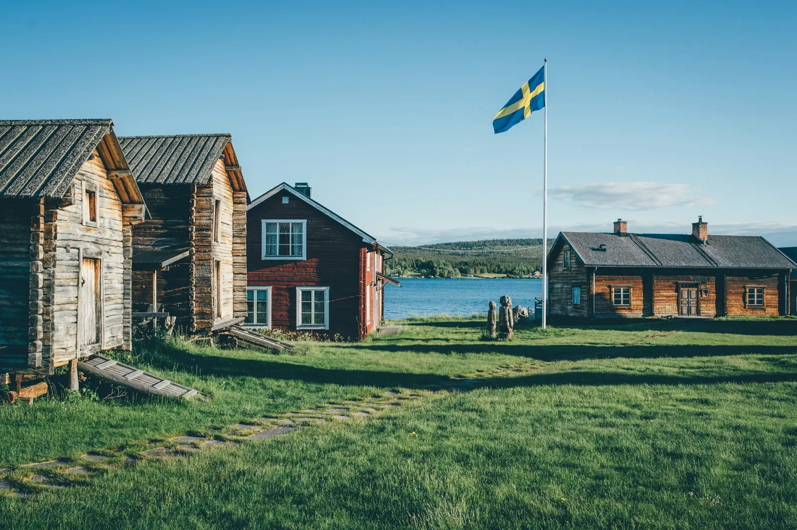 The Old Homestead in summer: weathered log buildings and a red wooden house around a lawn, a Swedish flag on its pole and the Torne River behind.