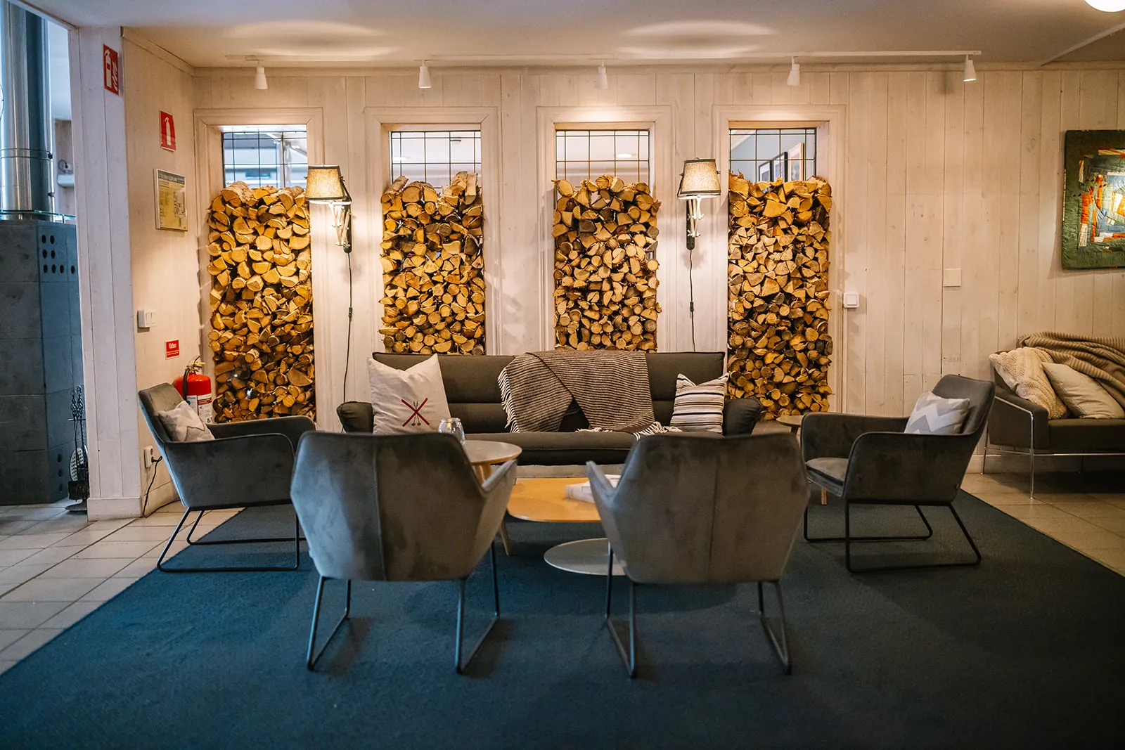 The lounge of Riverside Lobby: grey armchairs and a sofa on a dark carpet, pale wooden walls and 4 alcoves stacked with birch logs.