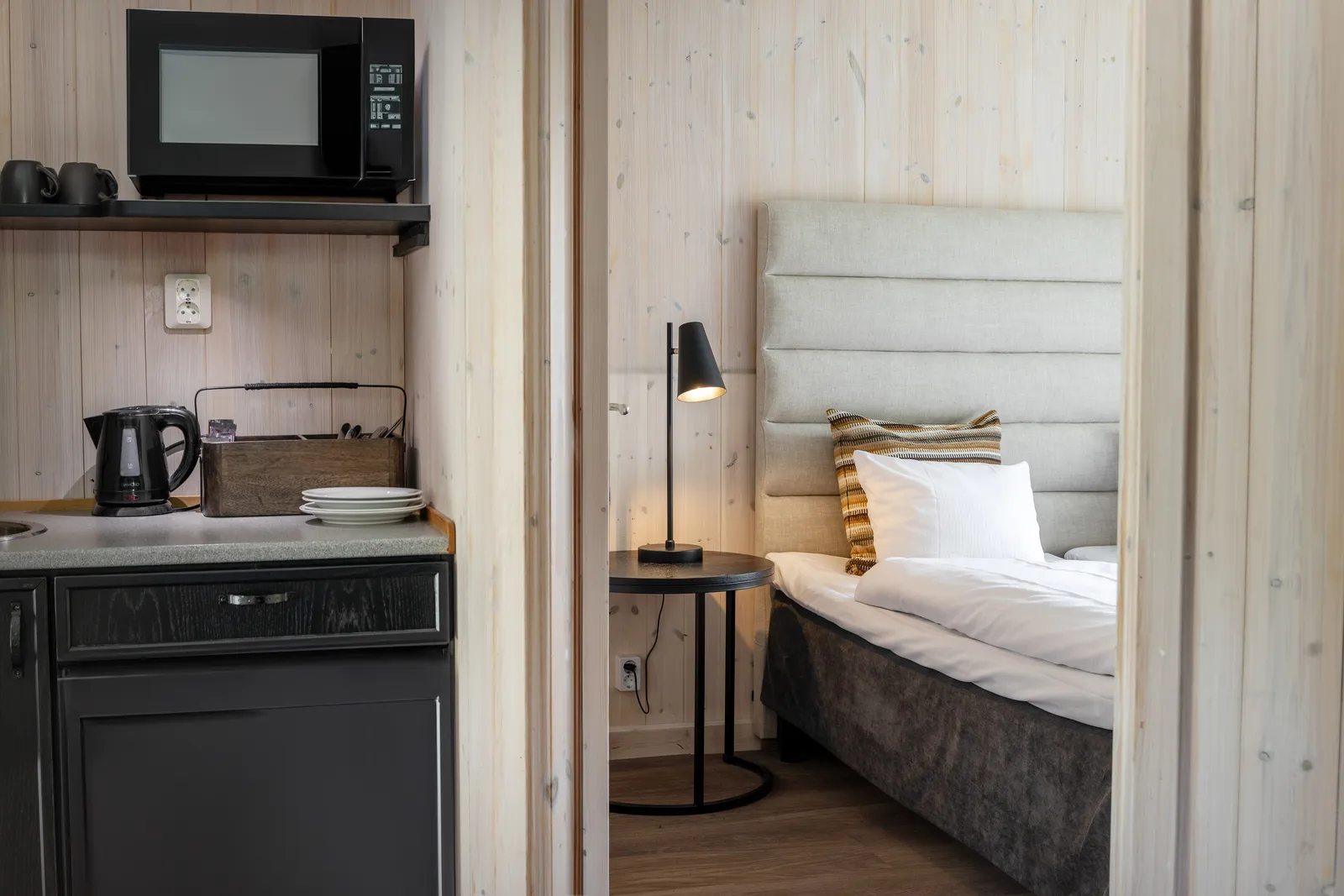 An Arctic Cabin: the kitchenette with microwave, kettle and fridge on the left and, through the doorway, a bed with an upholstered headboard and a reading lamp.