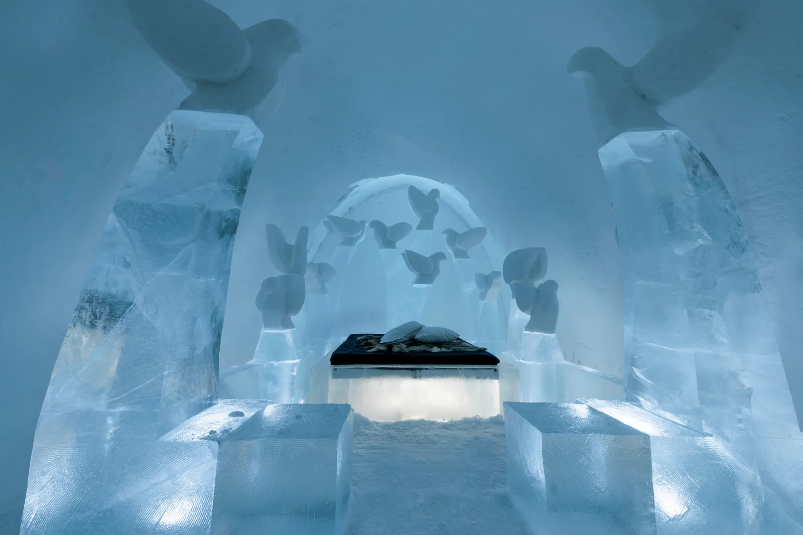 The Art Suite Flight, ICEHOTEL 35: a bed on a lit ice plinth beneath an arch of carved birds, between 2 ice pillars that end in wings.