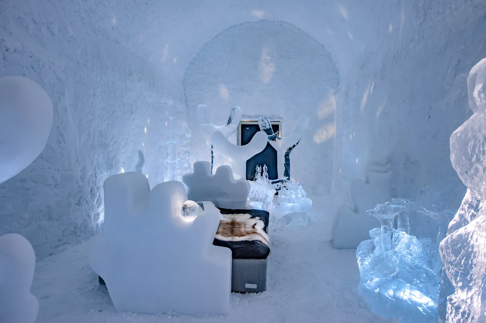 A Deluxe Suite in ICEHOTEL 365: a bed with a reindeer hide among rounded snow sculptures and blocks of clear ice, a dark doorway at the back of the room.