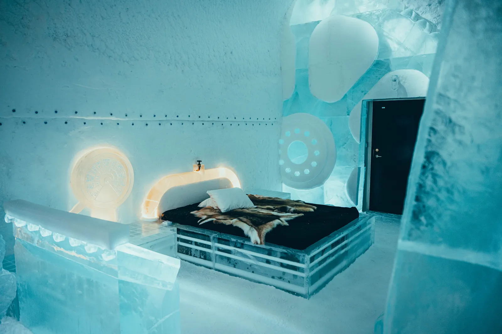 The Deluxe Suite The Breach: a bed on an ice frame with reindeer hides, a glowing ice headboard, ice portholes on the wall and the black door to the heated bathroom.