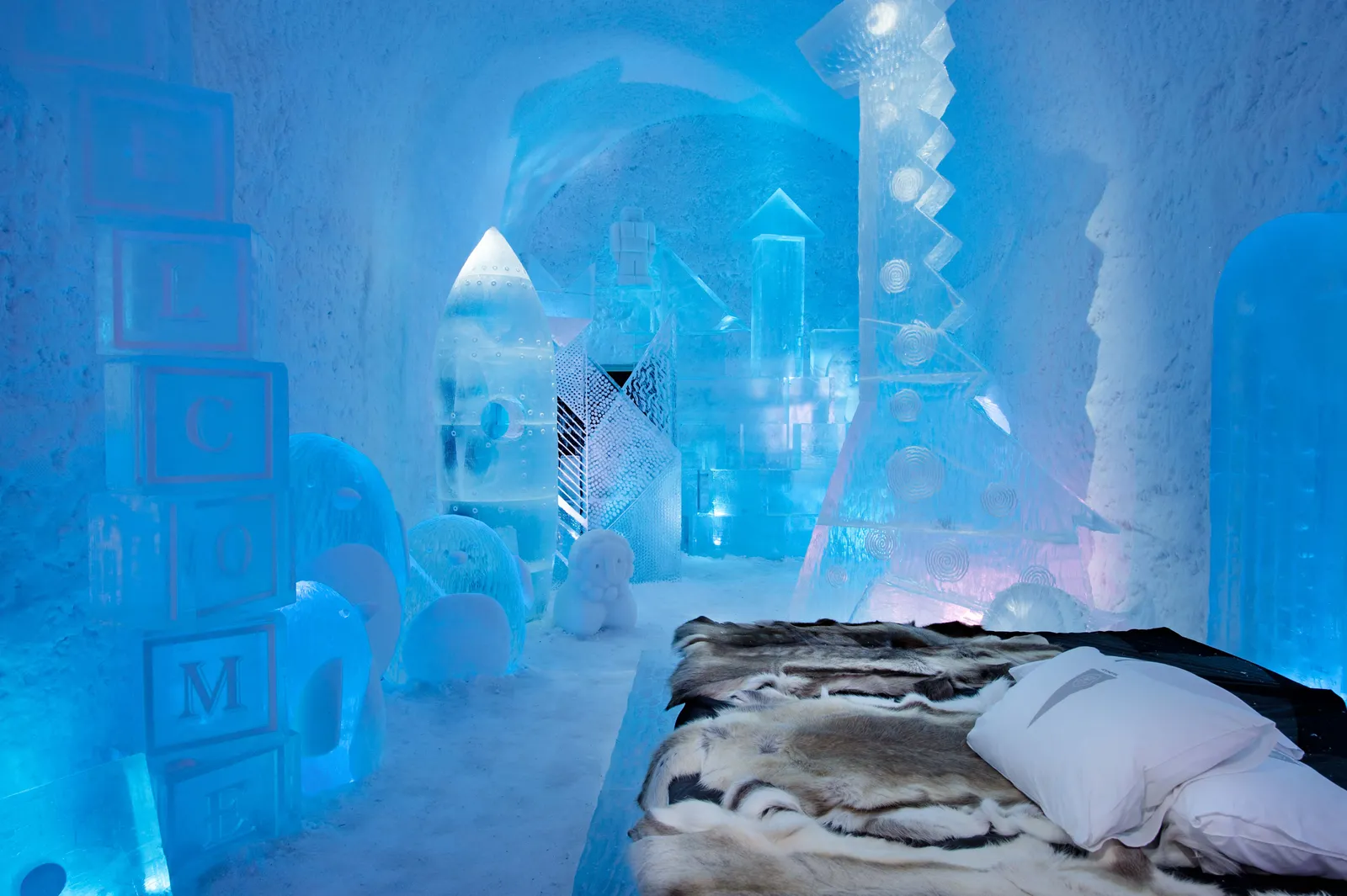The Deluxe Suite Toybox in ICEHOTEL 365: a bed with reindeer hides and white pillows, ice cubes lettered like toy bricks, a rocket and other toys carved in ice, in blue and pink light.