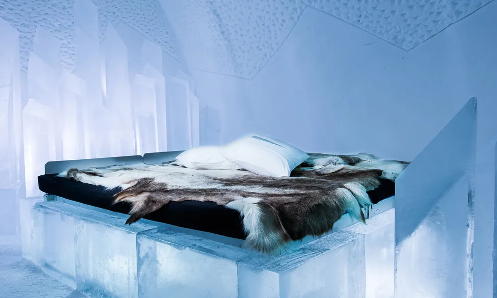An Ice Room: a bed on a base of ice blocks, covered with reindeer hides and 2 white pillows, between walls of snow and tall slabs of ice.