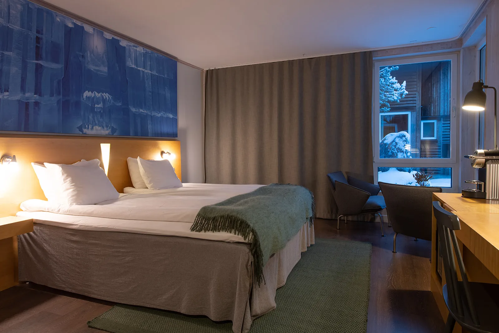 A Kaamos Deluxe room: a double bed with a green throw under a blue photograph of ice, armchairs by a window onto snow-covered houses, a desk with a coffee machine.
