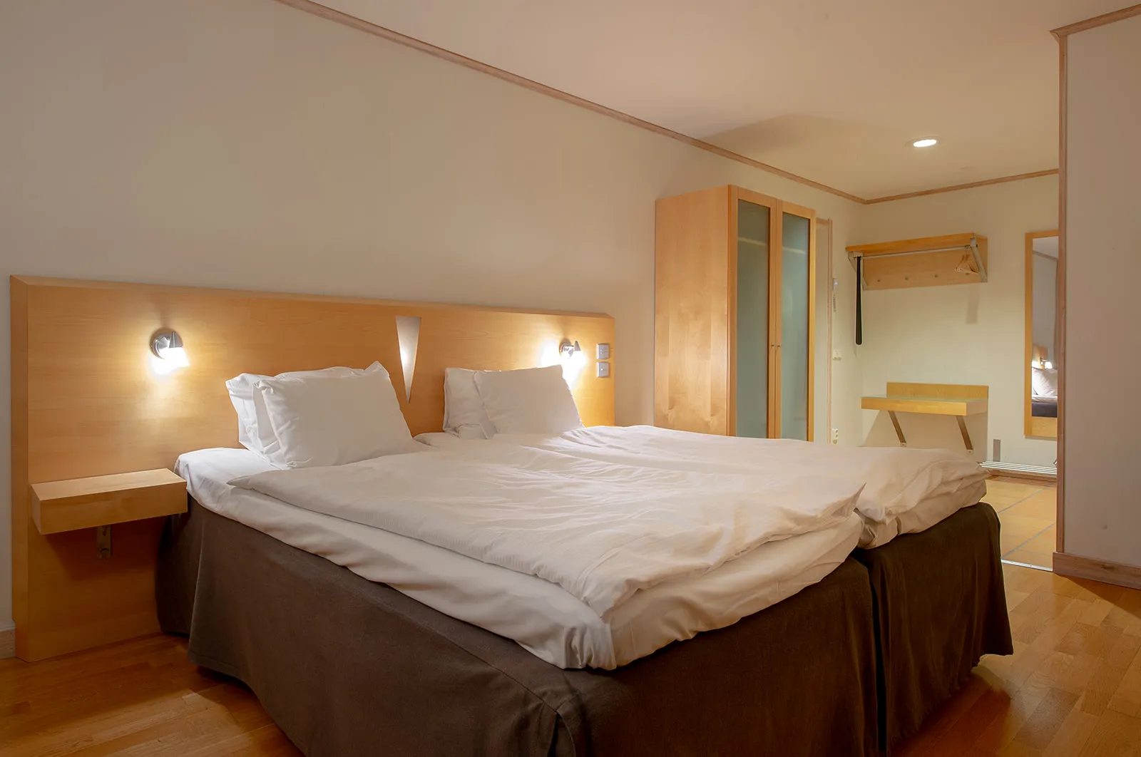 A Kaamos Hotel Room: twin beds pushed together with white duvets, a pale wooden headboard with 2 wall lamps, a wardrobe and a wooden floor.