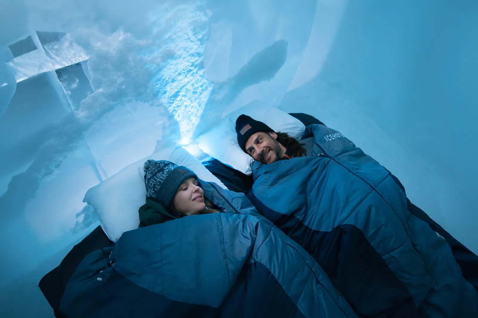 A couple in woollen hats lying side by side in blue sleeping bags on white pillows, under a vault of carved snow in blue light.