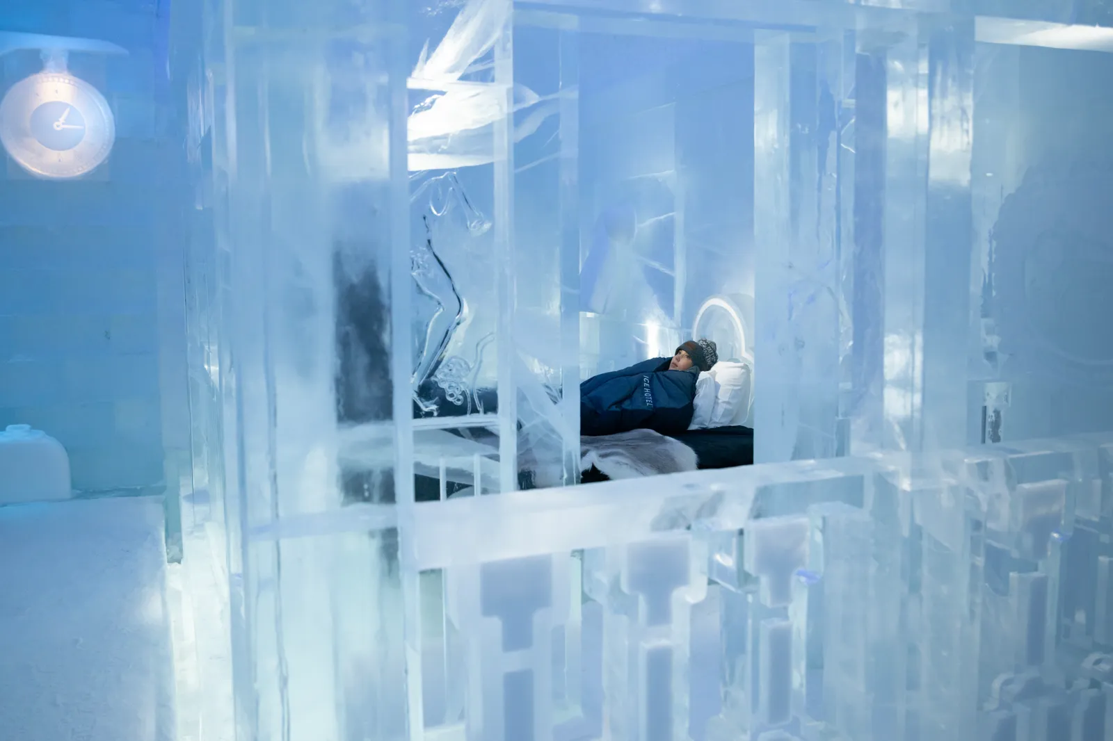 A guest in a hat lies in a blue sleeping bag on the bed of an ice suite, seen through carved panels of clear ice, with an ice clock on the wall.