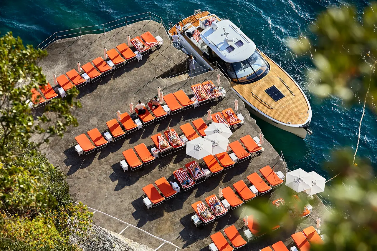 The beach club platform from above: rows of orange loungers on the rock and a wooden boat moored at the dock.