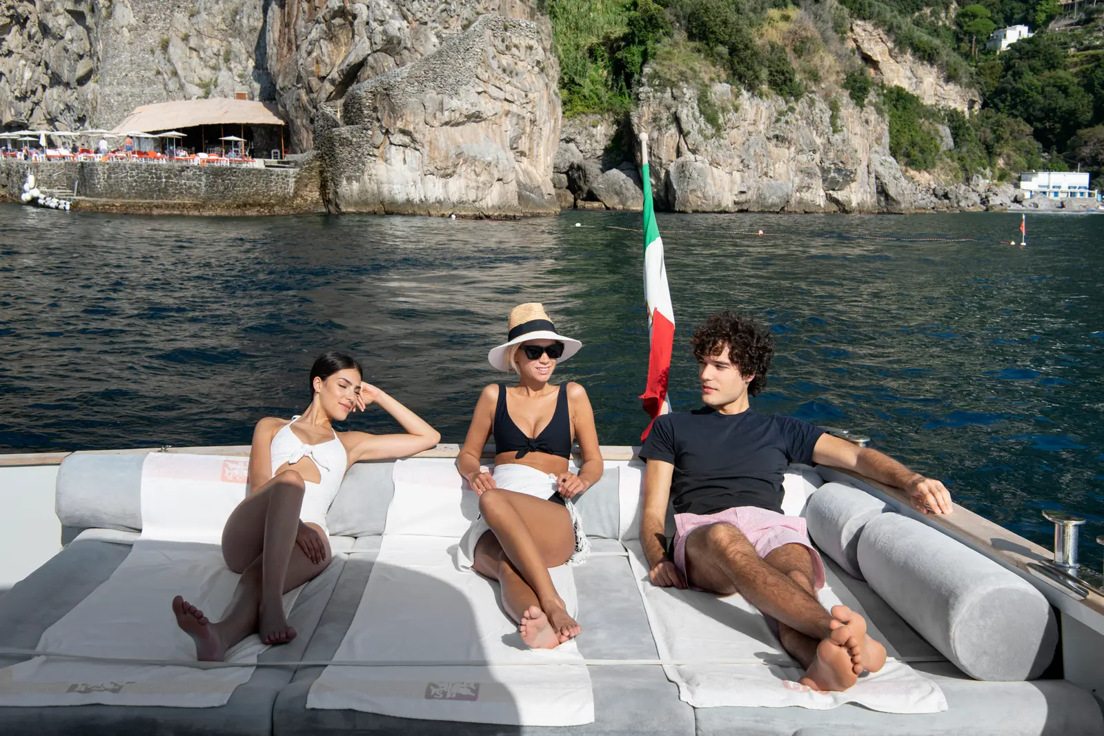 3 guests lying on the bow of the hotel’s boat, the Italian flag astern and the beach club’s cliff behind.