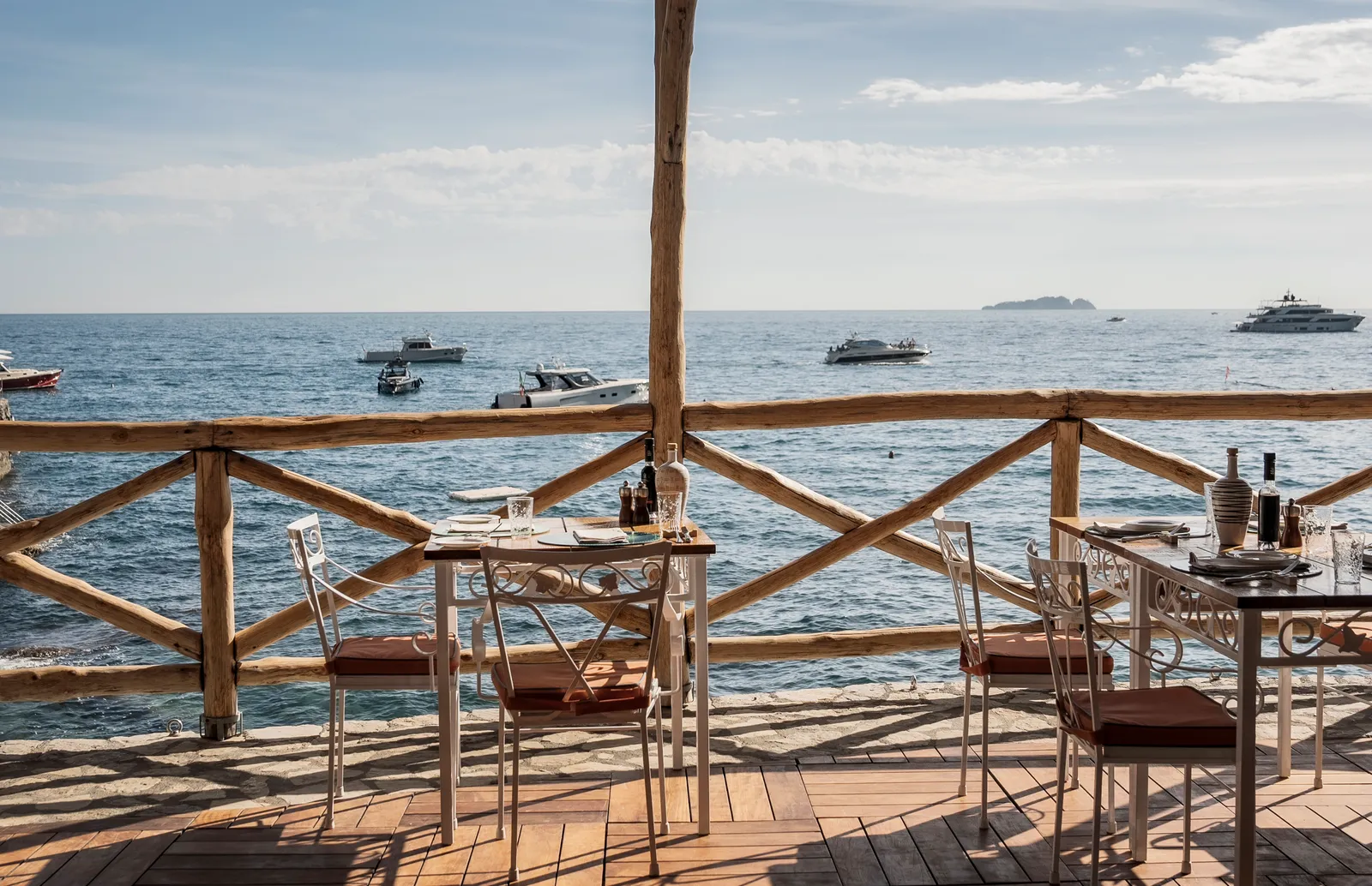 Carlino’s tables on the wooden deck at the water’s edge, behind a log railing, with boats at anchor.
