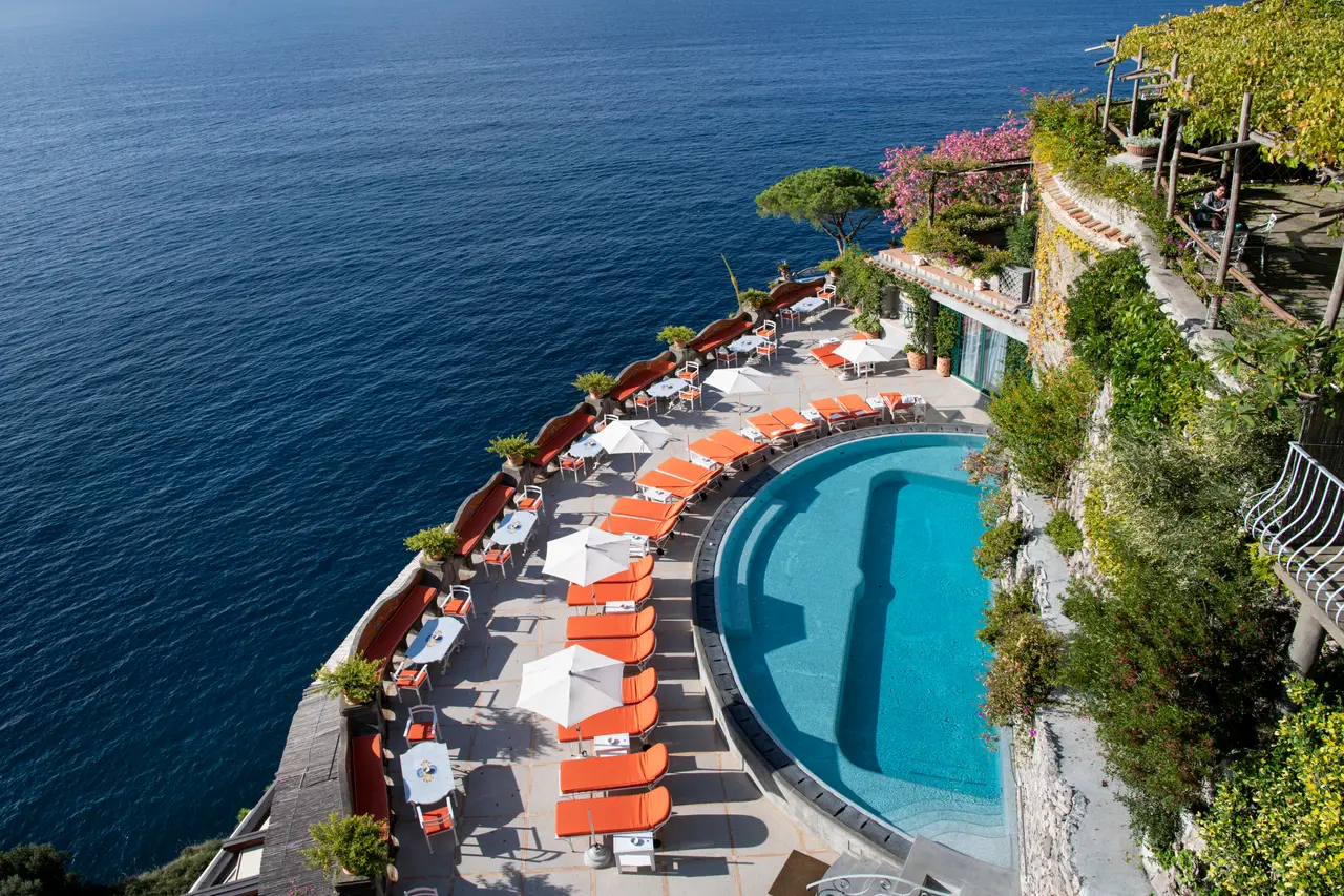 The crescent pool from above, with orange loungers and white parasols on the terrace sheer above the sea.