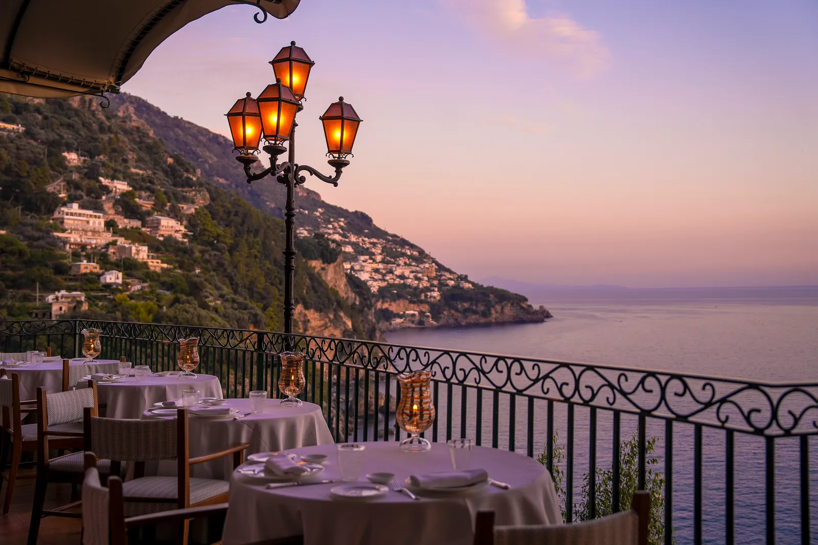 Laid tables on the restaurant terrace at sunset, a lit street lamp and the Positano coast under a pink sky.
