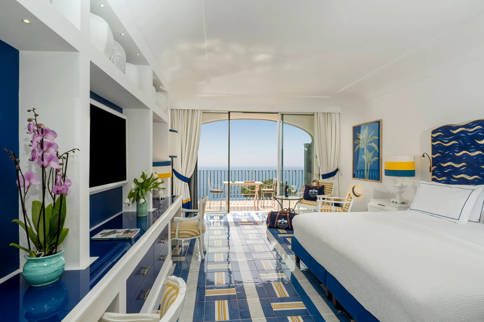 A long, bright Premier room, with a blue wave-patterned headboard, orchids and the arch open onto the terrace and the sea.