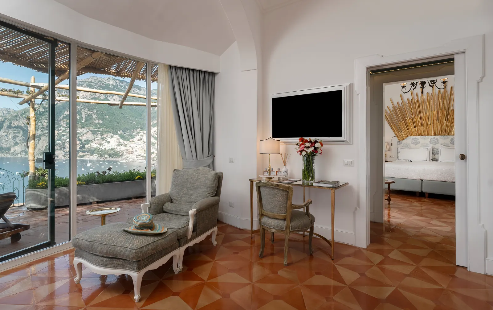 The sitting room of a Prestige with Positano view: terracotta floor, a grey chaise longue and the window onto the pergola terrace.