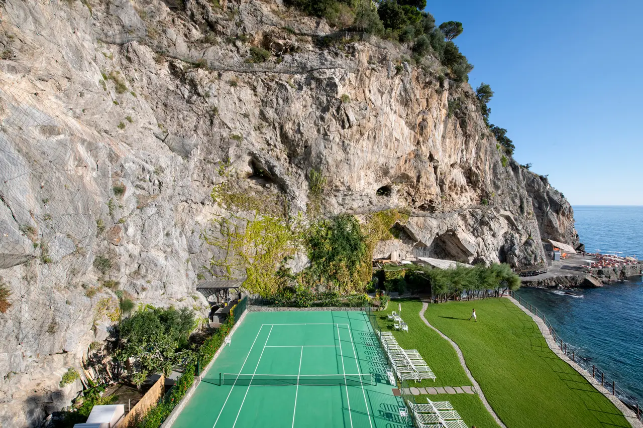 The green tennis court at the foot of the rock face, beside the lawn and the sea, with the beach club beyond.