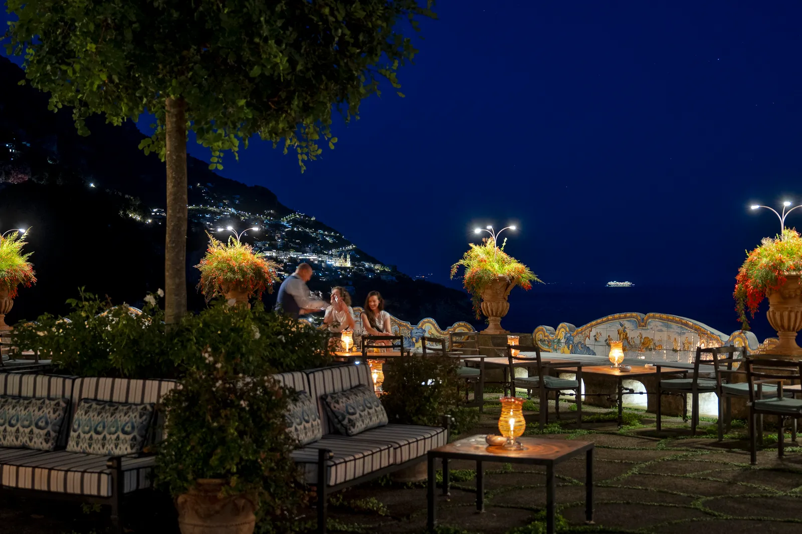 The Terrace Bar at night: striped sofas, lit urns of flowers and the majolica benches before the lights of Positano.