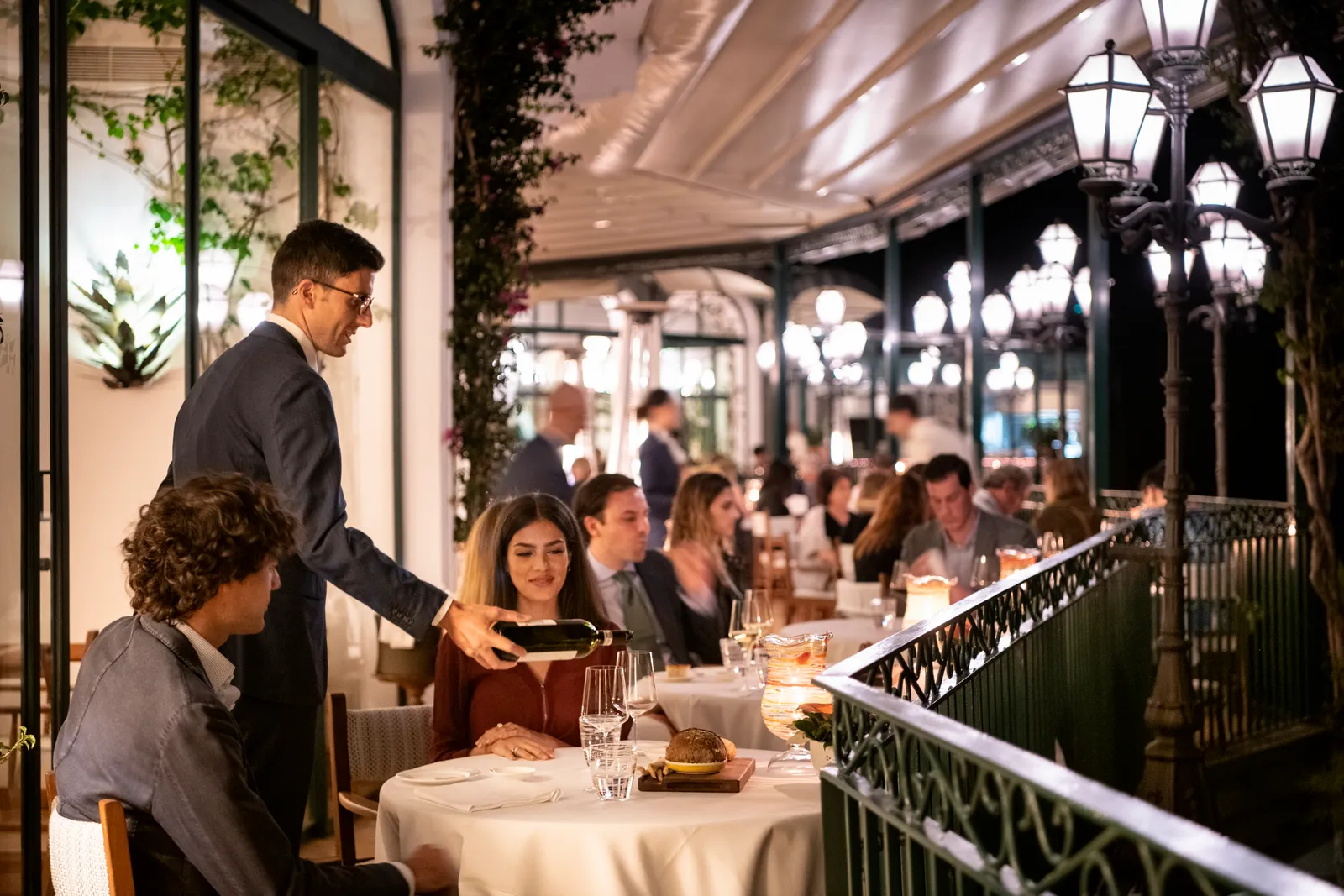 Dinner at Zass: a waiter pours wine at a table for 2 on the veranda, the full room behind and the lamps lit.