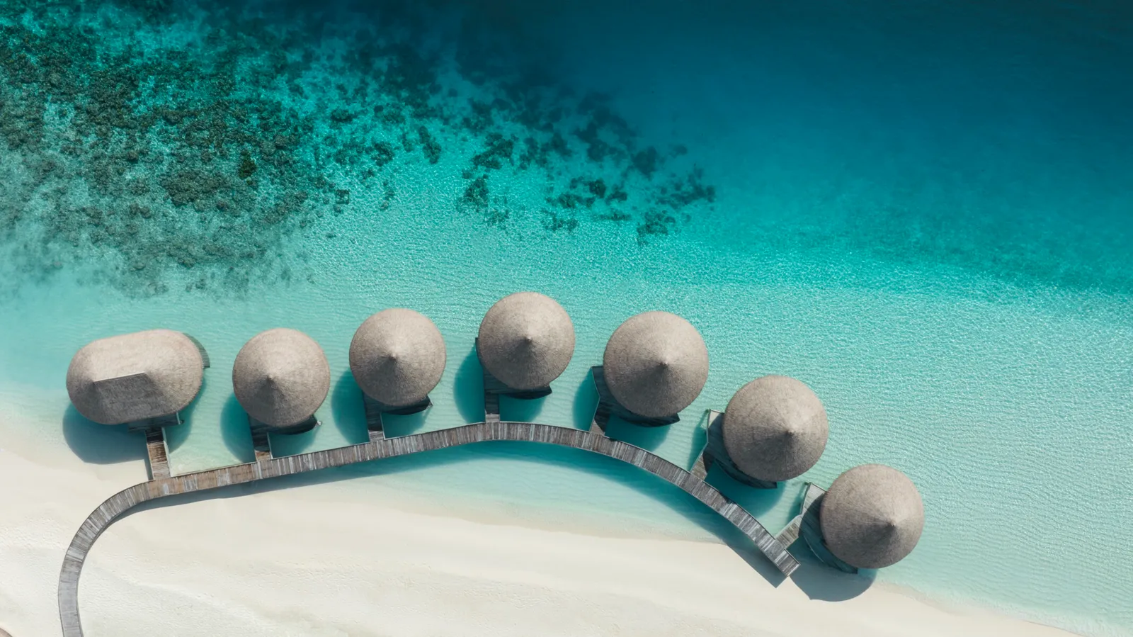 AVI Spa’s treatment villas from above: round thatched roofs along a curving jetty over the lagoon.