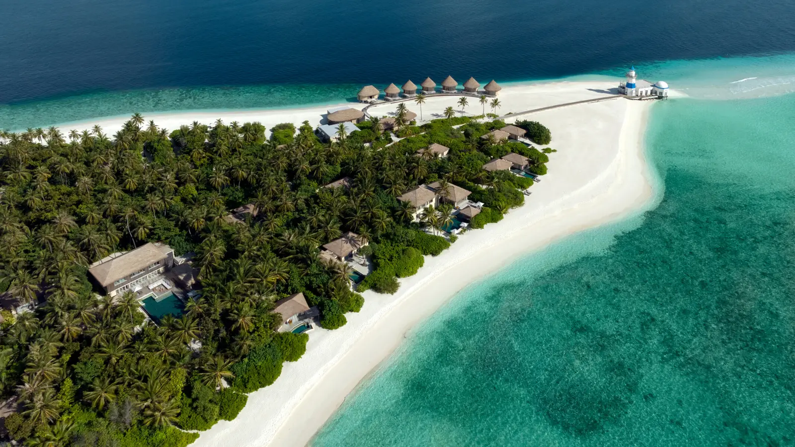The island from above: villas among the palms, the white spit of sand, the row of water villas and the lighthouse at the tip.