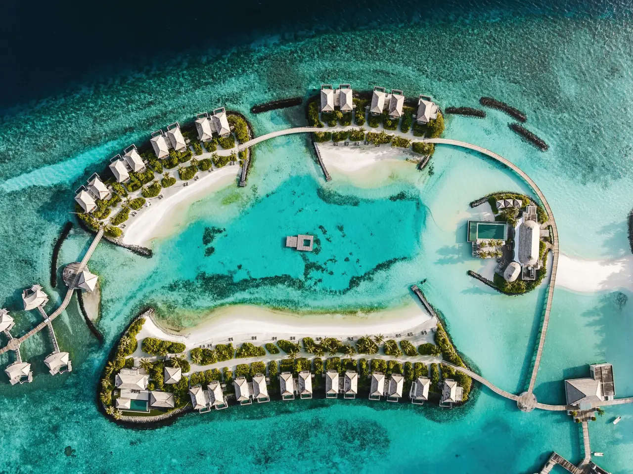 The lagoon held by the island, from above: beach villas among the palms and water villas along the curving jetty.