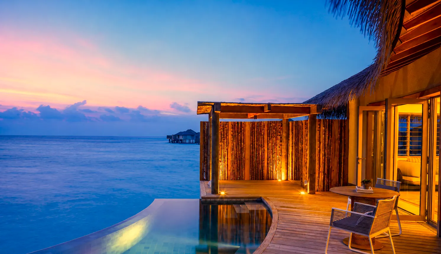 A Sunset Overwater Pool Villa at dusk, the deck lit and the infinity pool facing west.
