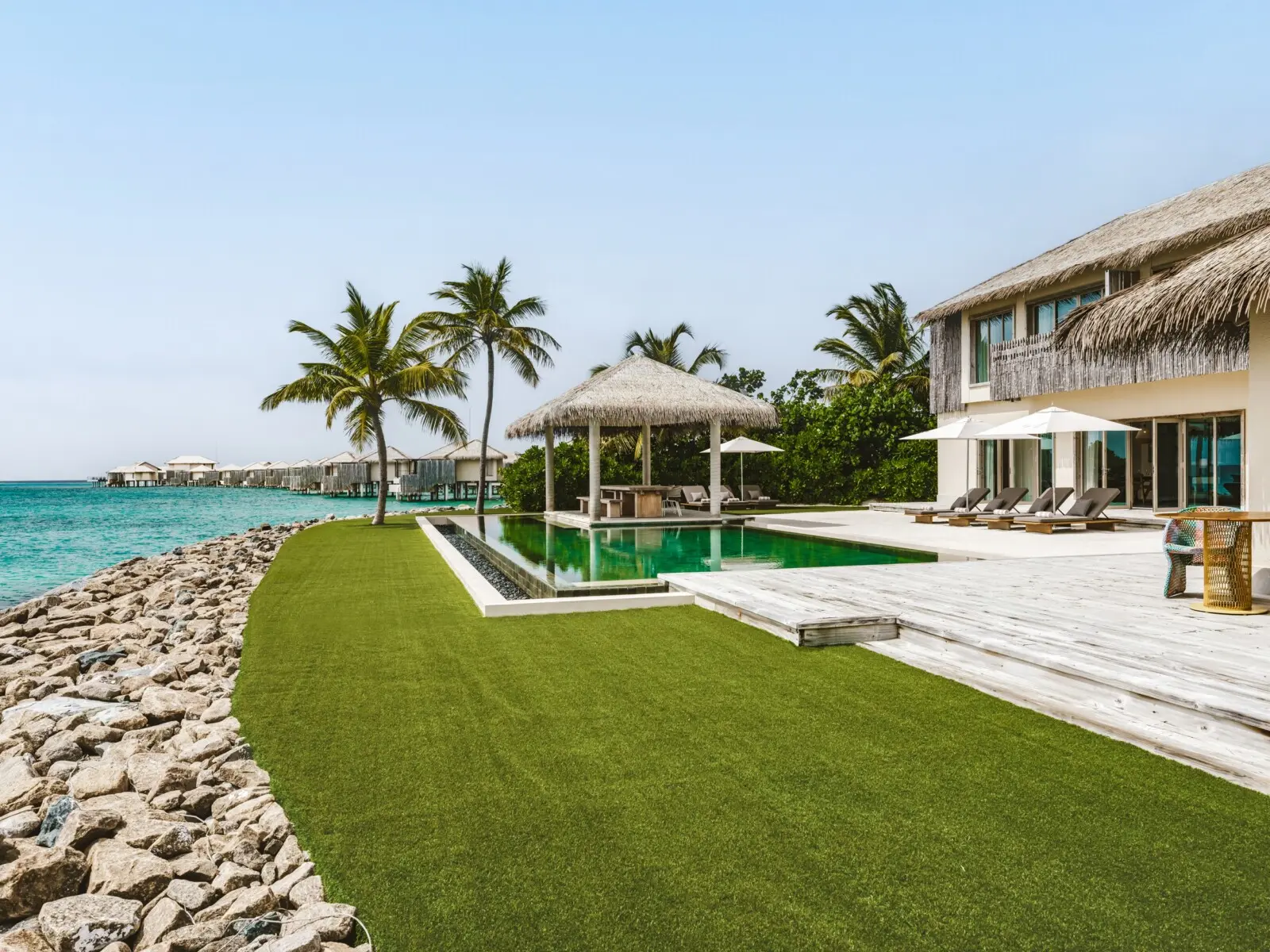 The Three Bedroom Lagoon Pool Residence’s lawn, the long pool with its thatched gazebo and the sea beyond the rocks.