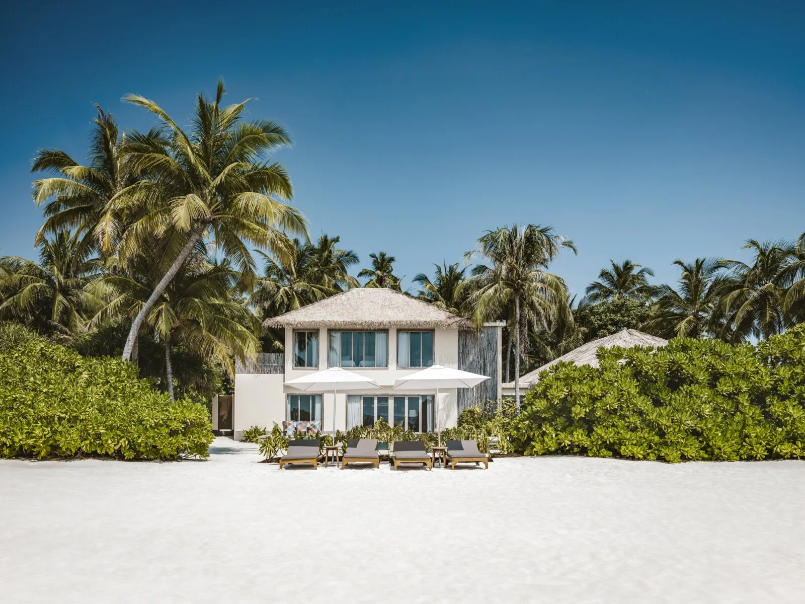 The Two Bedroom Beachfront Pool Residence, a white 2-storey house among the palms, loungers on the sand.