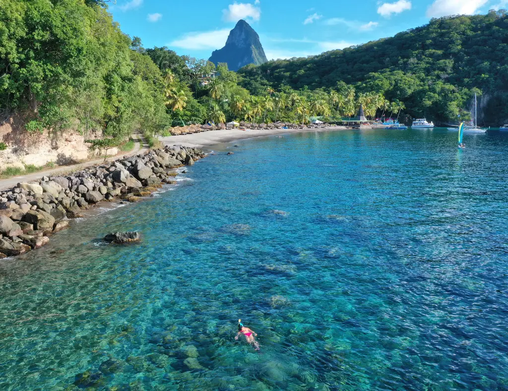 A snorkeller in clear turquoise water off Anse Chastanet beach, with the rock breakwater, the palms and the tip of Petit Piton.