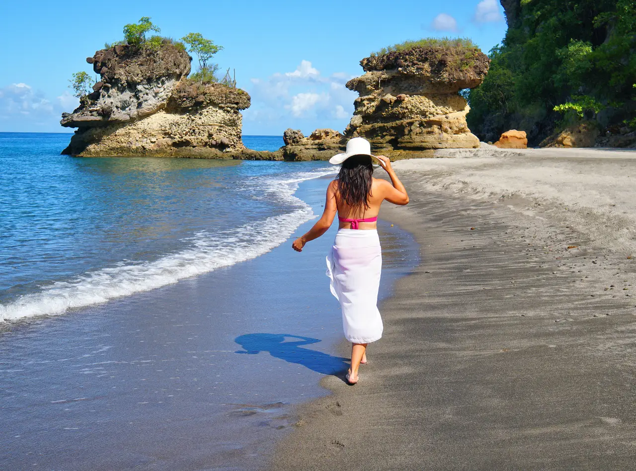 A woman in a white hat walking along the grey sand of one of the 2 beaches, towards 2 rock stacks topped with greenery.