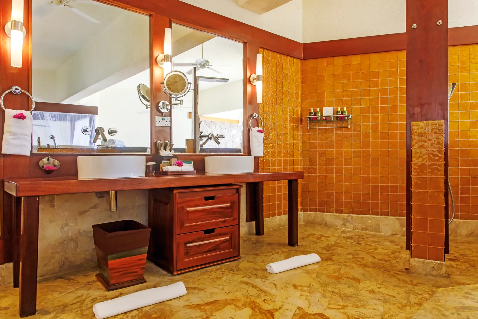 A sanctuary’s open bathroom: 2 basins on a timber counter, mirrors, a wall of amber glass tiles and a marble floor.