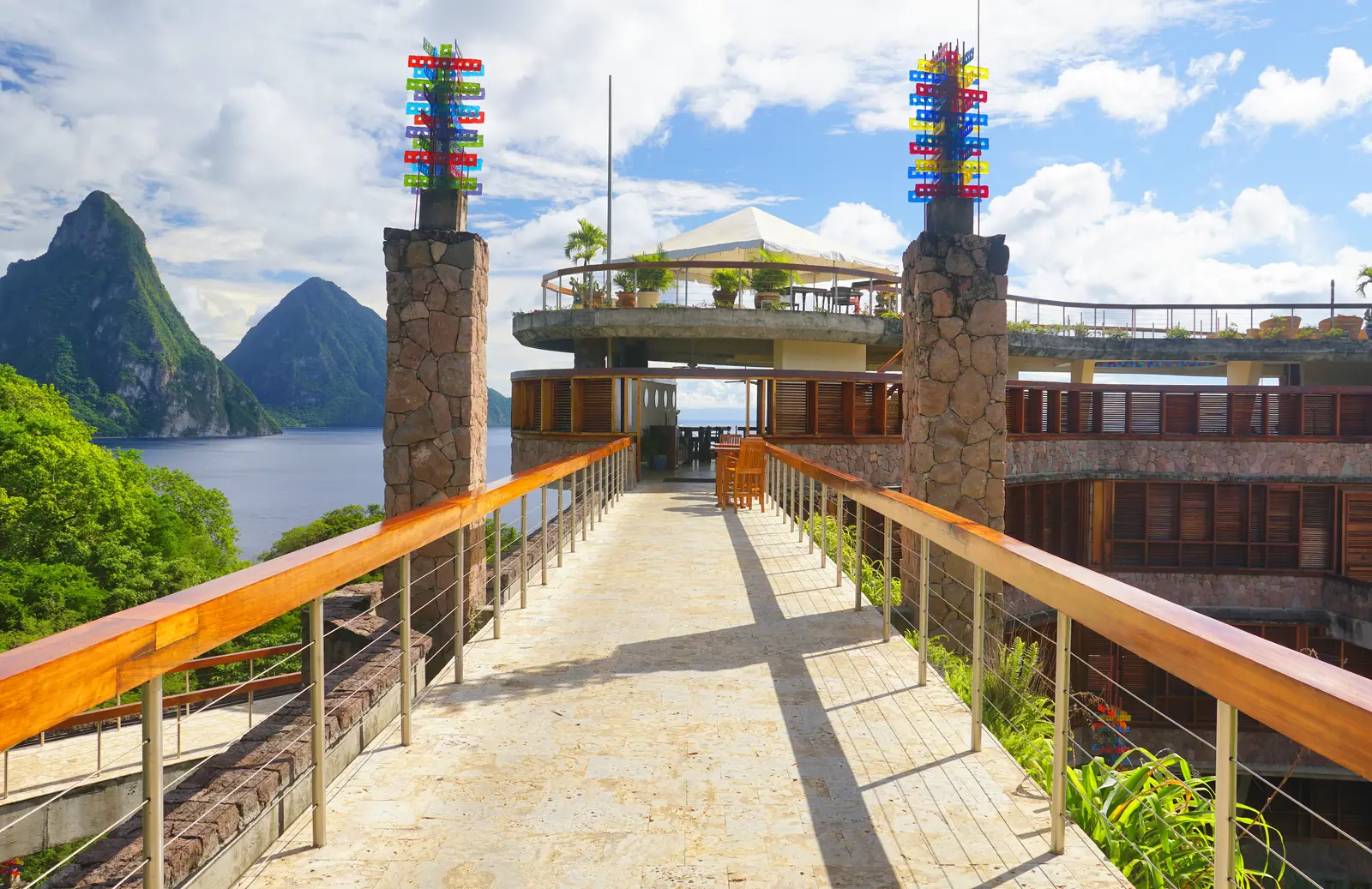 One of the footbridges to the building: its timber handrail, 2 stone pillars topped with coloured sculptures, the Celestial Terrace and the Pitons to the left.