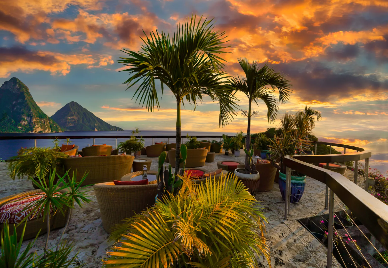 The Celestial Terrace at sunset: wicker armchairs among potted palms, the rail, an orange sky and the Pitons in silhouette.
