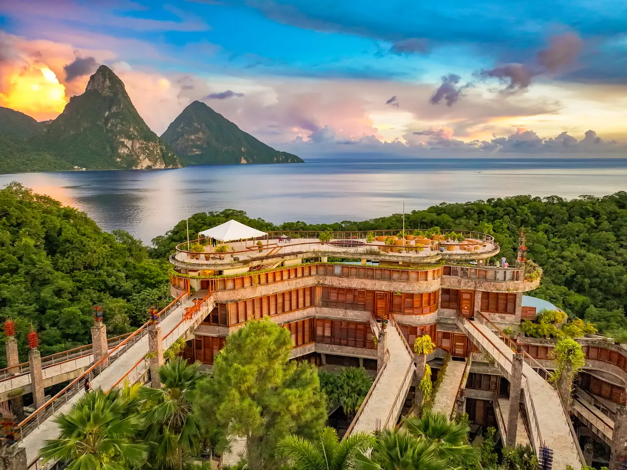 Jade Mountain from above at sunset: its stepped floors and the suspended bridges leading to the sanctuaries, forest all round, the bay and the 2 Pitons beyond.