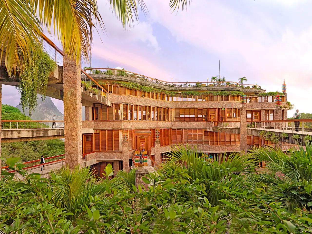 The building’s inner face: 4 floors of stone and timber, the footbridges reaching them, a palm in the foreground and tropical planting.