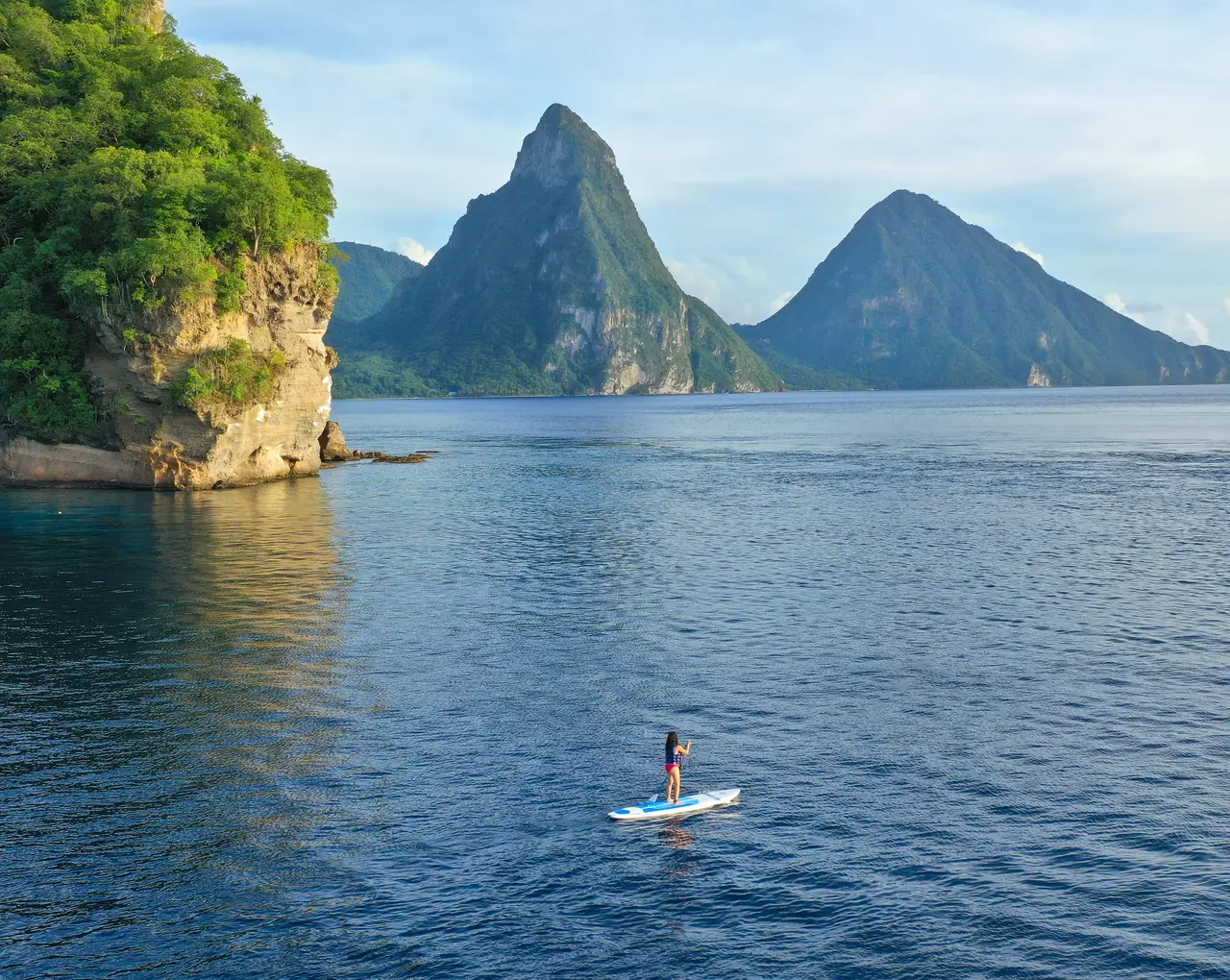 A paddleboarder on calm water under the cliff, with the 2 Pitons behind.