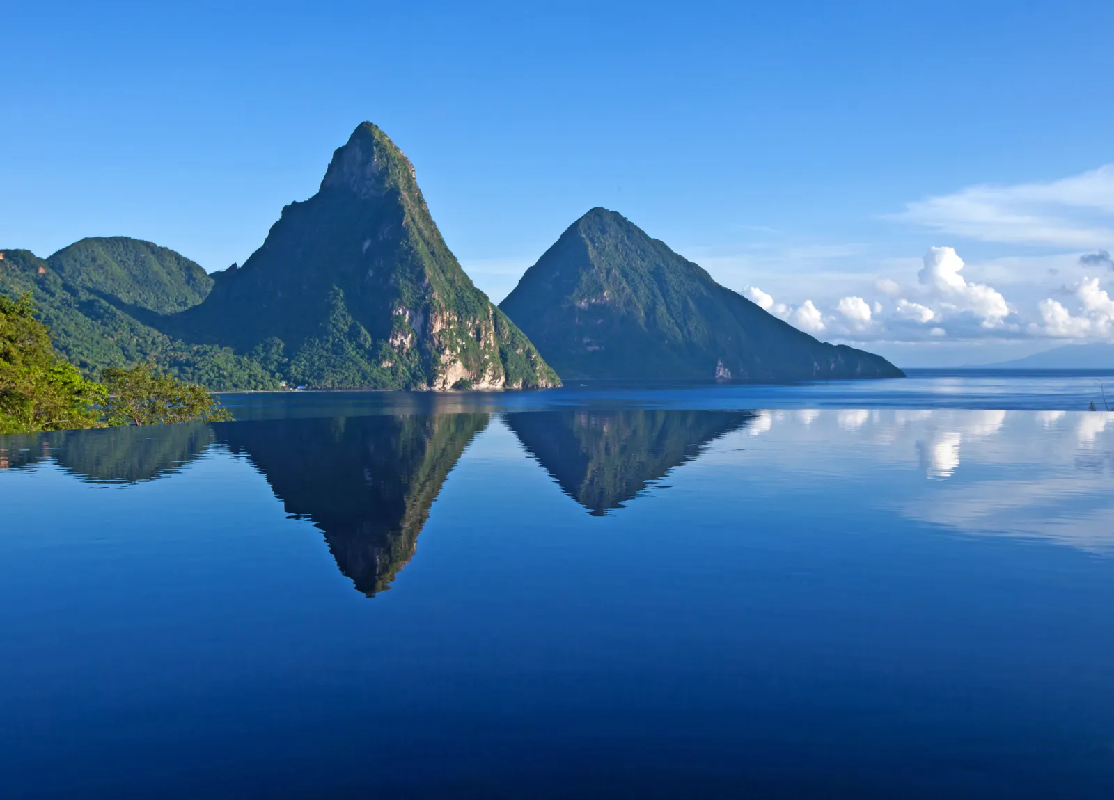 The still water of an infinity pool mirroring the 2 Pitons, with the sea and a blue sky beyond its edge.