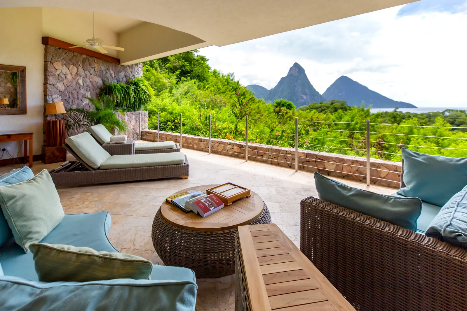 A Sky suite: 2 loungers and a wicker sitting area with pale blue cushions on the room’s open side, a steel-cable rail, the forest and the Pitons.